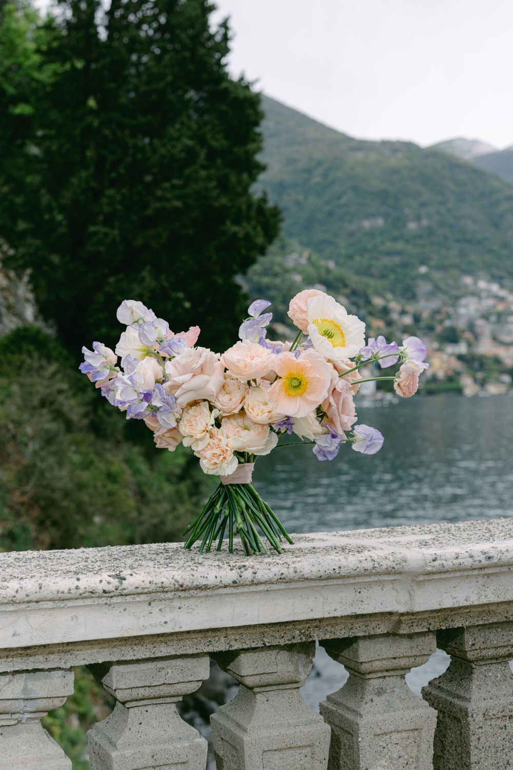 Delicate bouquet of pastel flowers arranged on stone balustrade overlooking a lake and mountains.