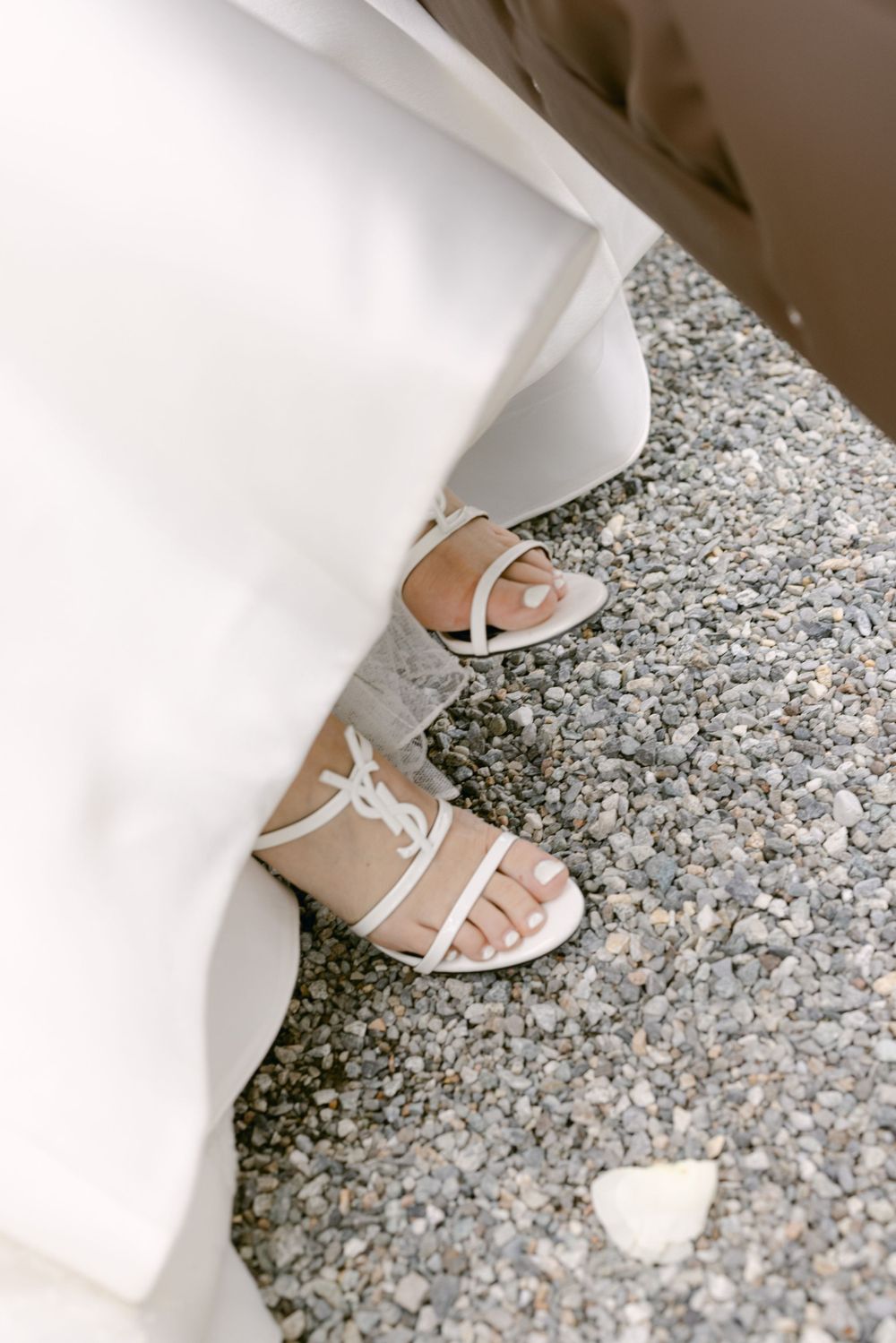 White strappy sandals peeking out from beneath a flowing white dress on gravel ground.