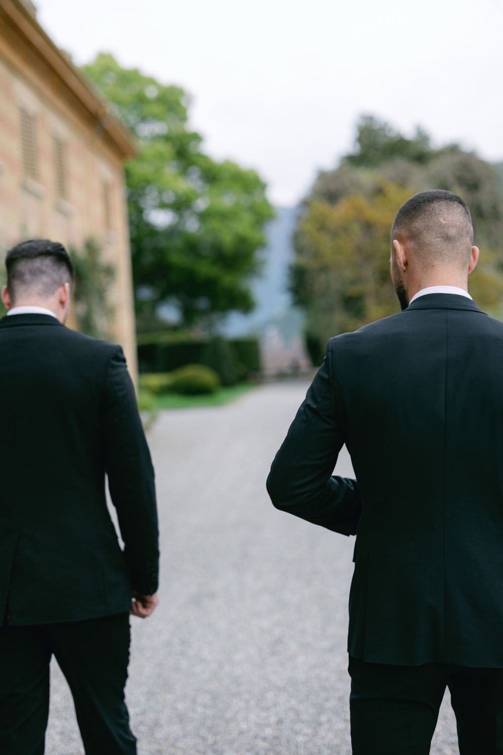 Two men in black suits walking down a gravel path from behind.