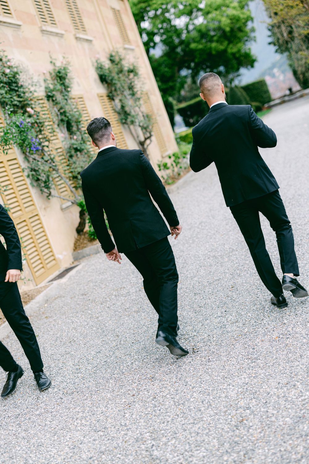 Three people in black suits walking down a gravel path near a pink building.