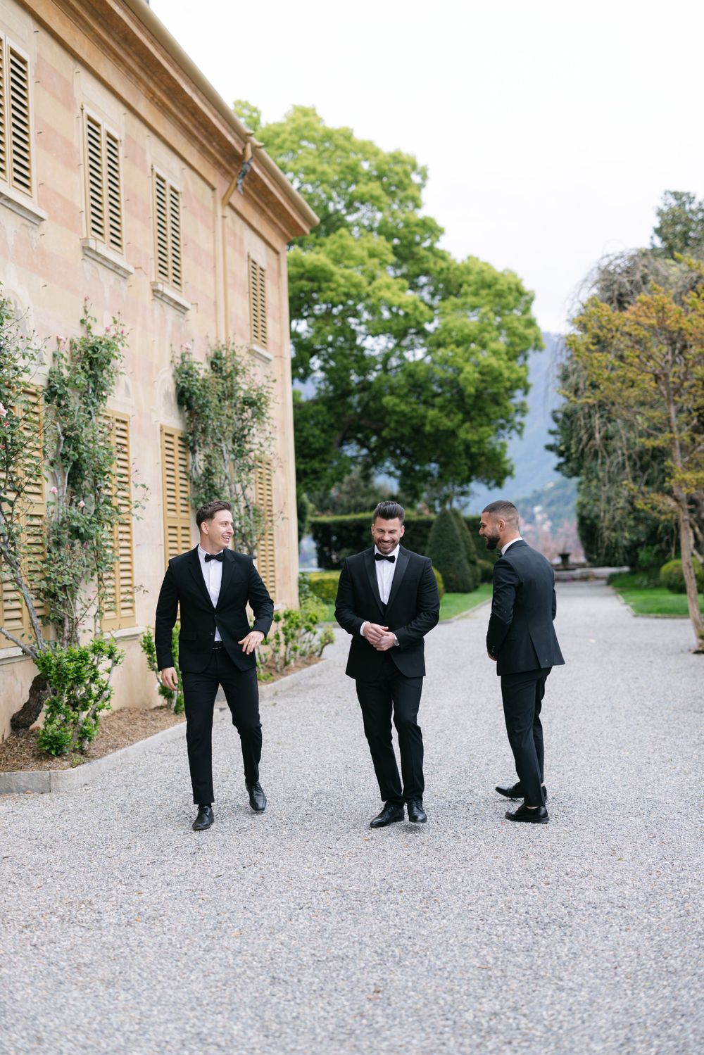Three people in formal attire walking along a gravel path beside a Mediterranean-style building.