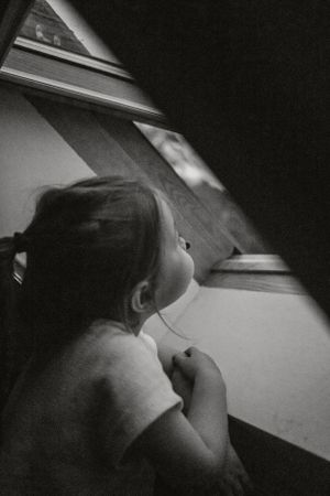 Black and white image of child looking up through window in dramatic lighting.