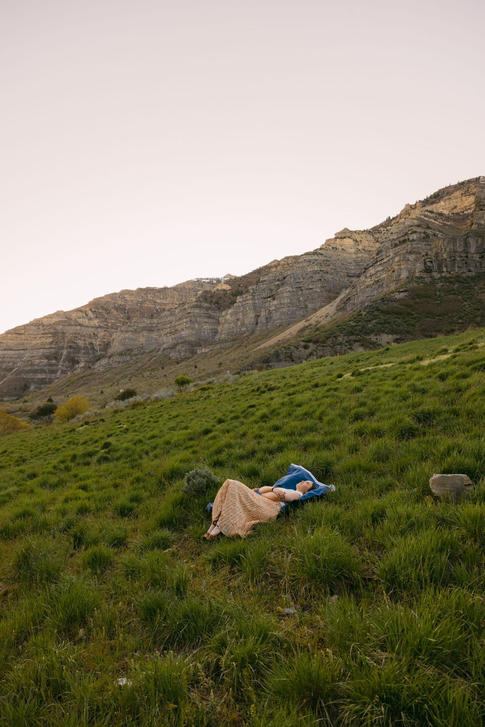 Person lying in green mountain meadow with dramatic rocky cliffs in the background.