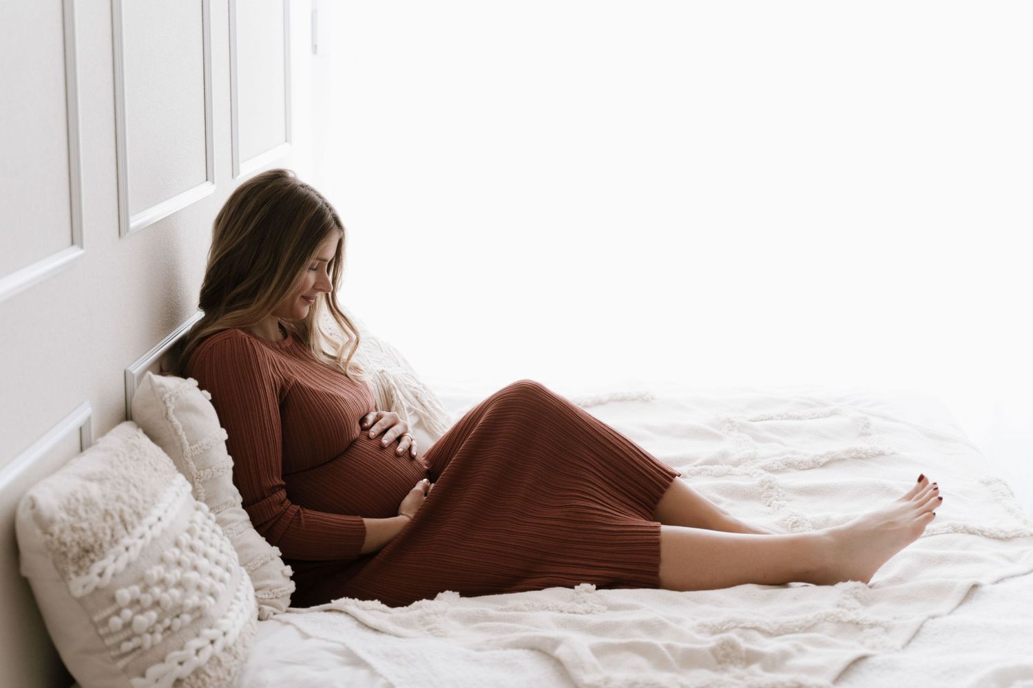 Pregnant woman in a rust-colored knit dress sitting on a white bed, gently cradling her baby bump.