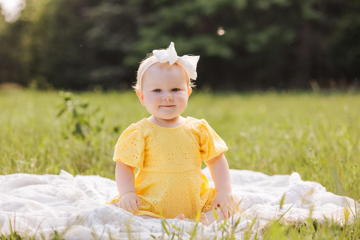A baby in a yellow outfit sits on a white blanket in a sunny meadow.