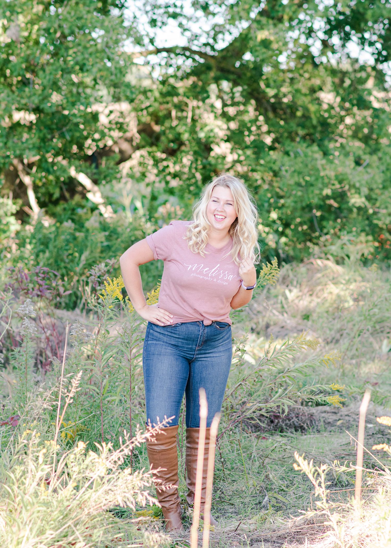 Smiling blonde woman in a mauve graphic tee and jeans posing outdoors among lush green foliage.