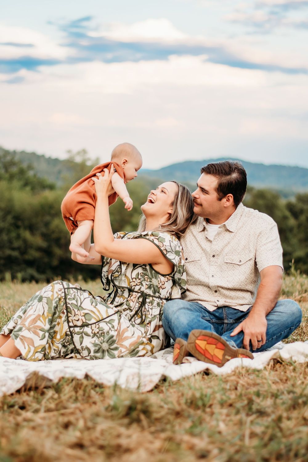 Laughing couple sitting on blanket in meadow lifting baby in rust outfit with mountains behind.
