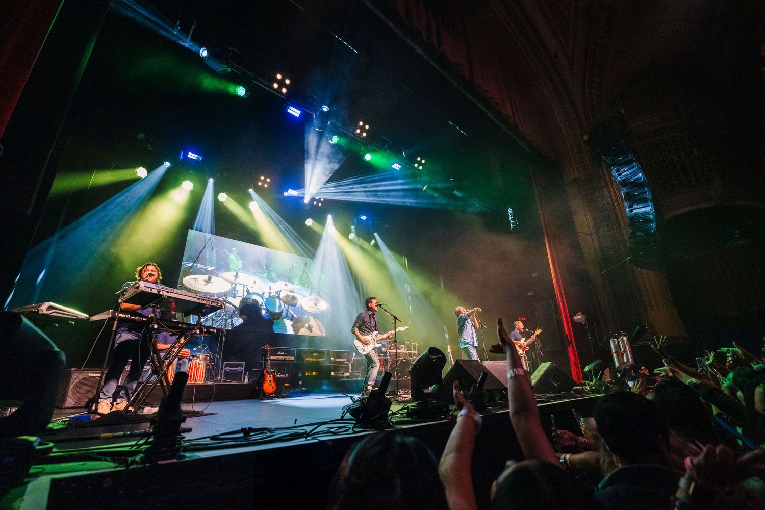 Concert stage with multiple performers and dramatic light show at packed indoor venue.