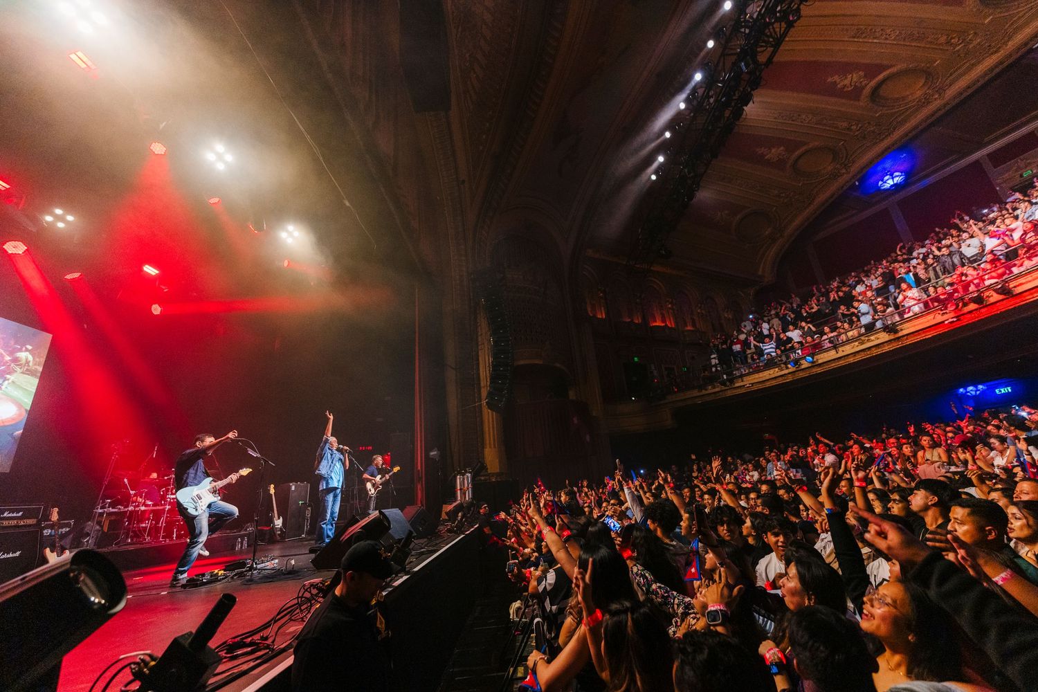 Wide shot of crowded theater with red lighting and performers on stage during live concert.