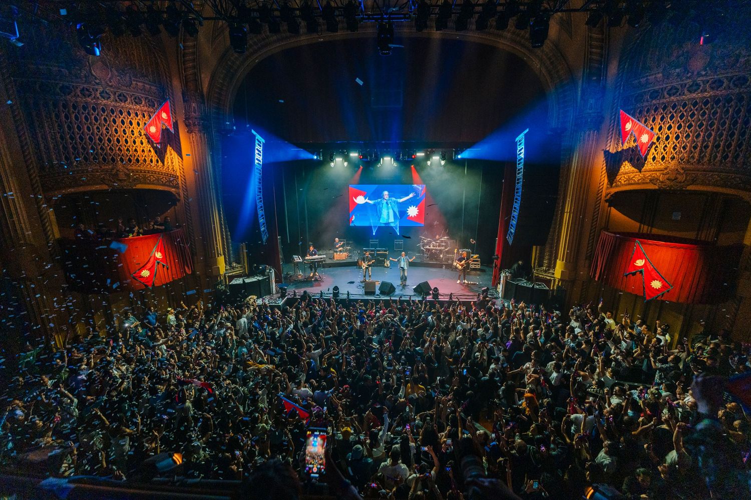 Aerial view of packed theater showing crowd and performers on stage with dramatic blue and red lighting.