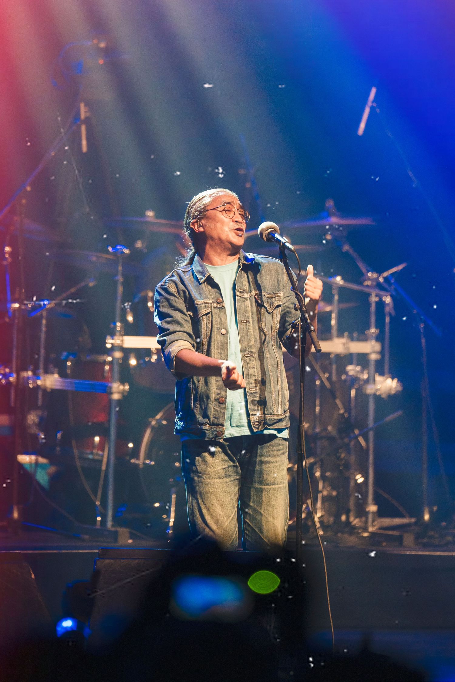 Vocalist performs on concert stage with dramatic blue lighting and drum kit visible in background.