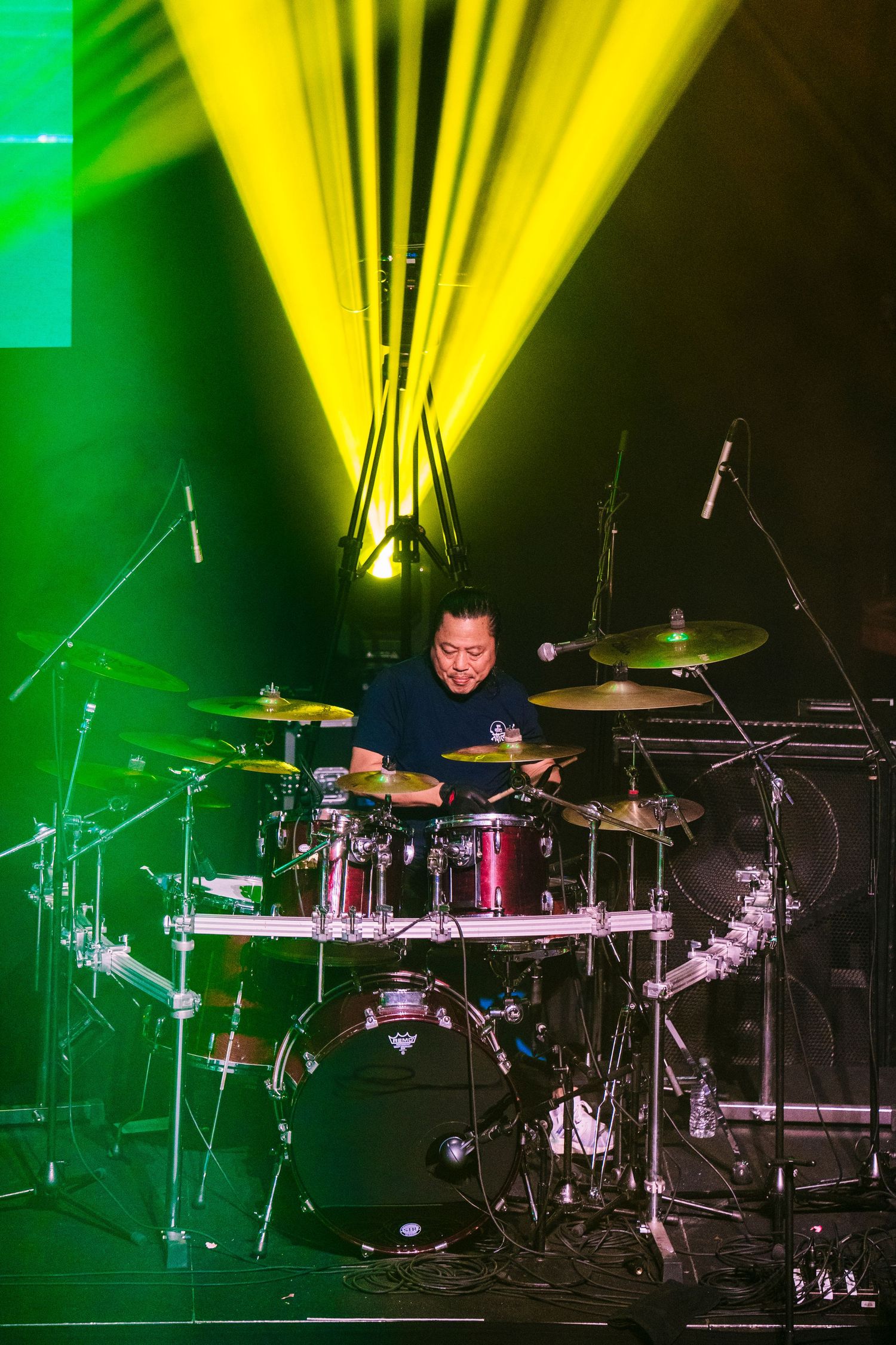 Drummer performs on elevated drum kit platform with dramatic yellow and green stage lighting.