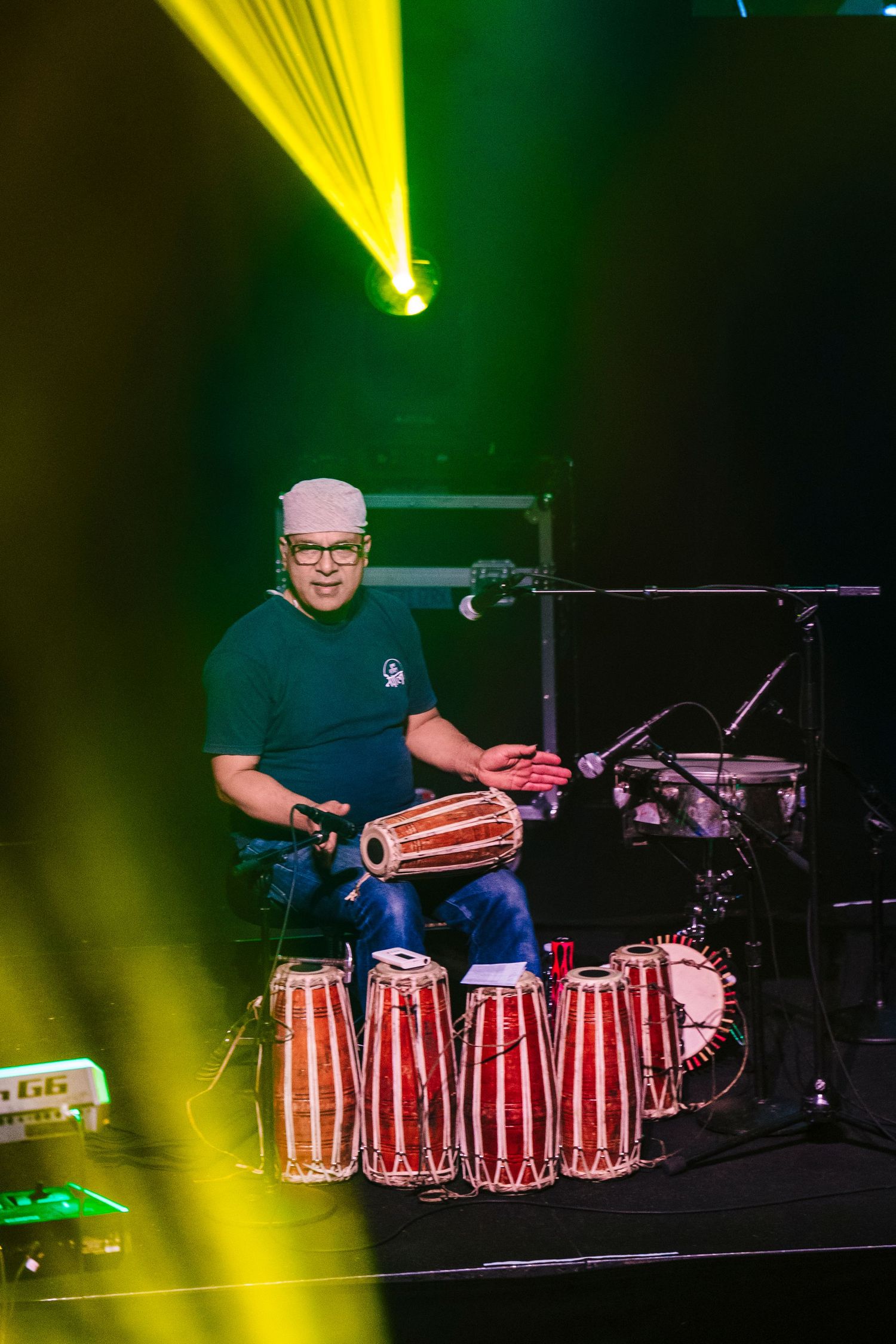 Percussionist plays multiple conga drums under dramatic yellow and green stage lighting.