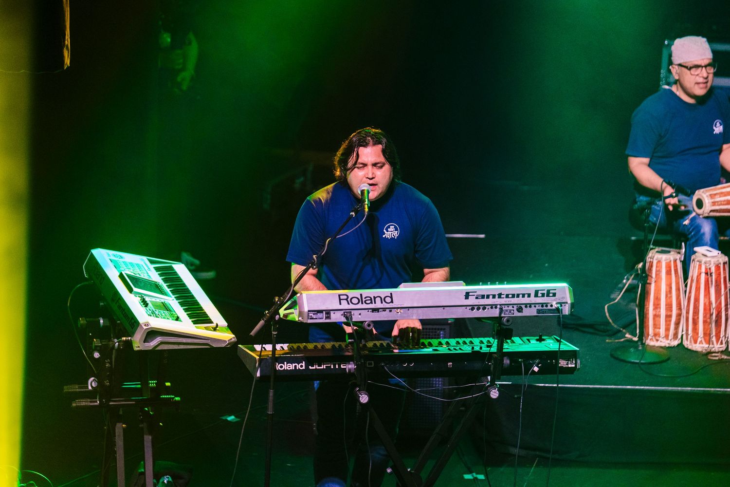 Keyboardist performs on Roland synthesizer while percussionist plays congas in background.