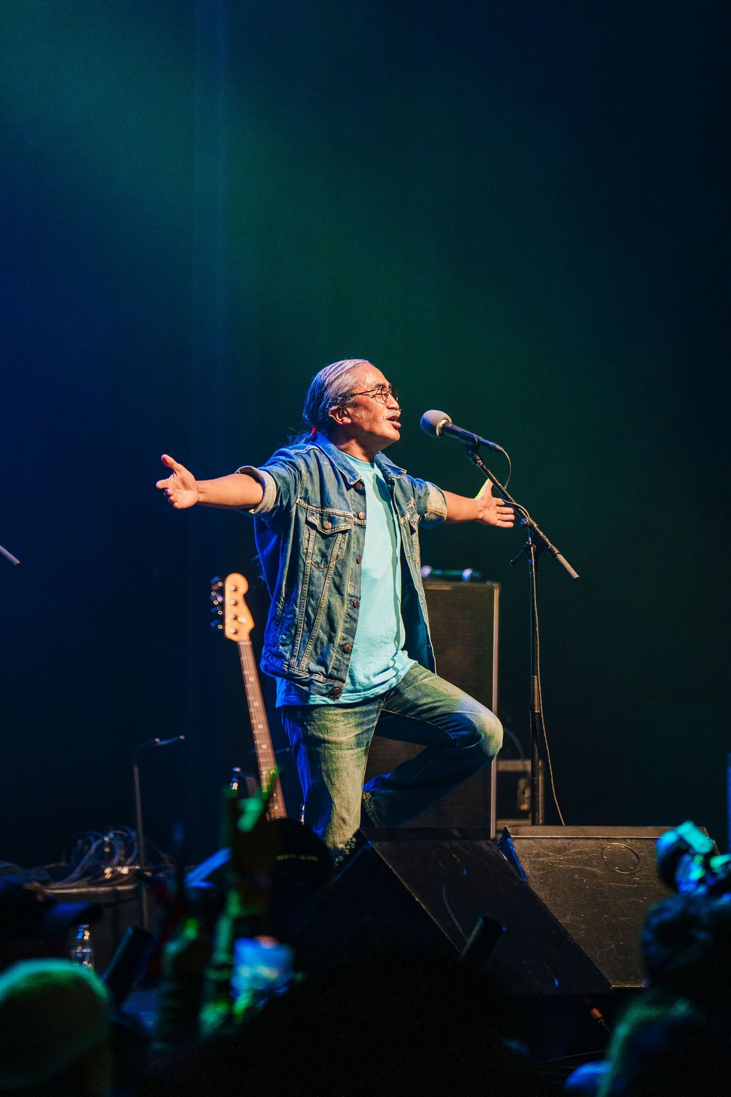 Energetic performer in denim vest sings passionately on stage with dramatic lighting and crowd in background.