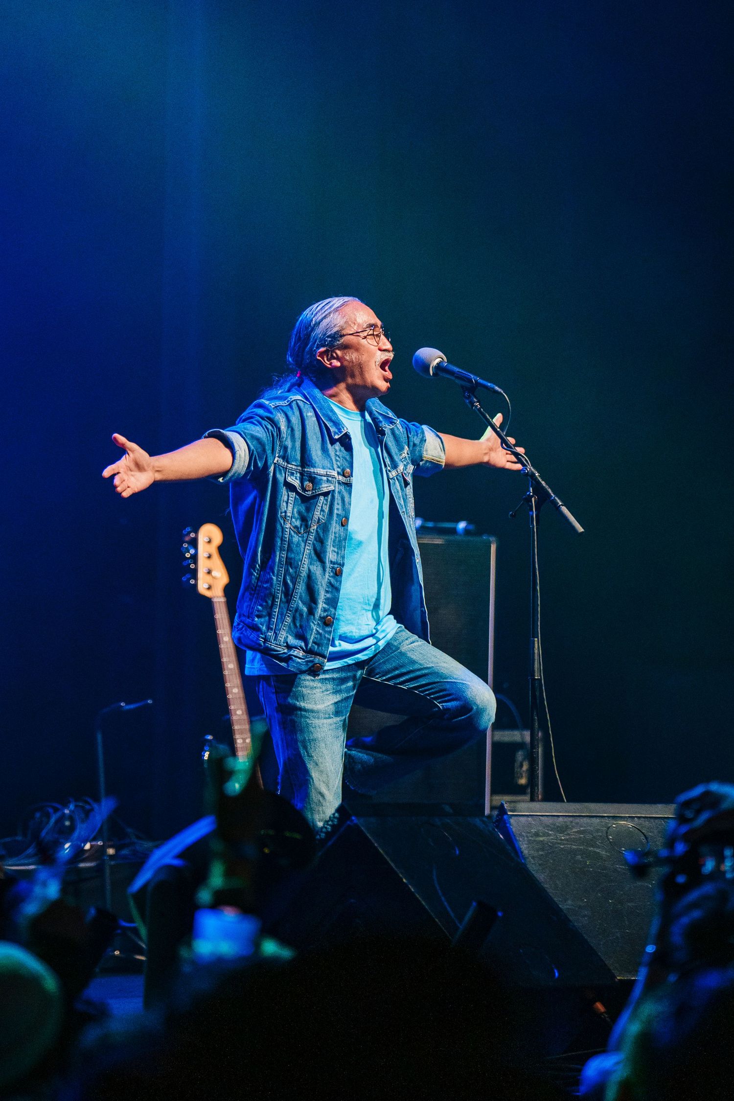 Singer spreads arms wide while performing energetically on concert stage with blue lighting effects.