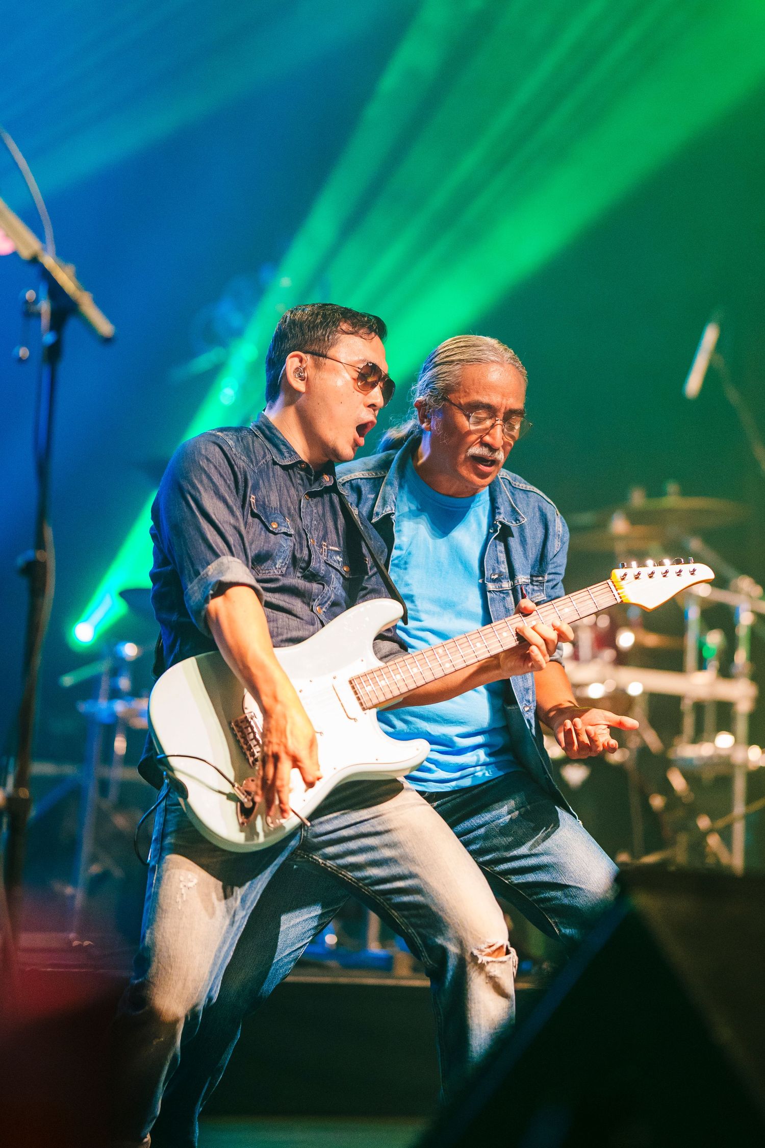 Two musicians perform together on stage with electric guitar under vibrant green and blue lighting.