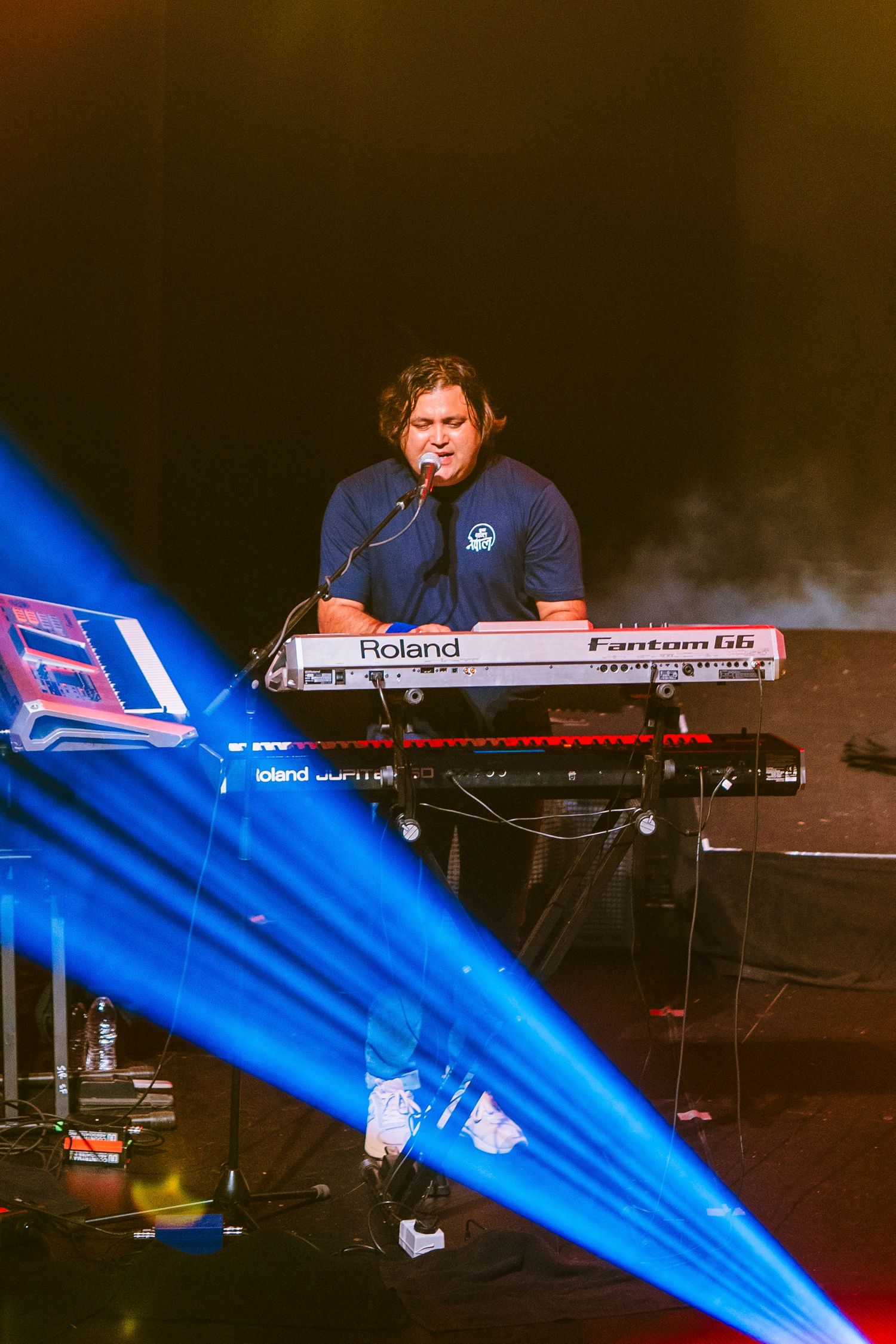 Musician plays Roland keyboard on stage with dramatic blue spotlight beam.