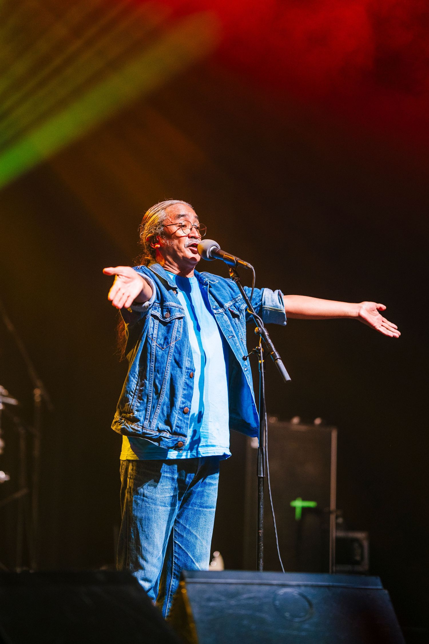 Performer in denim vest spreads arms wide during energetic stage performance with colorful lighting.