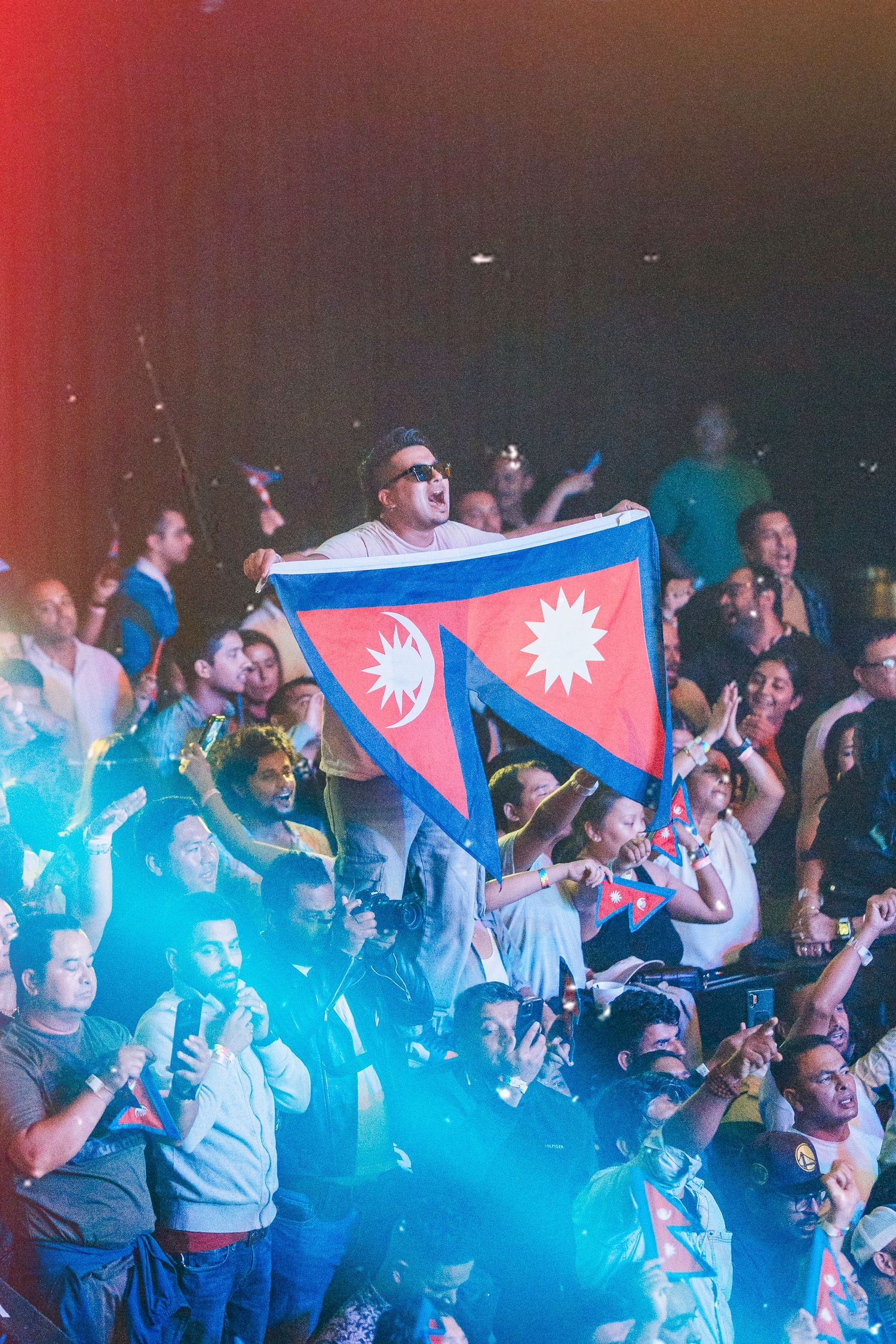 Concert crowd waves Nepali flag during energetic performance with blue lighting.
