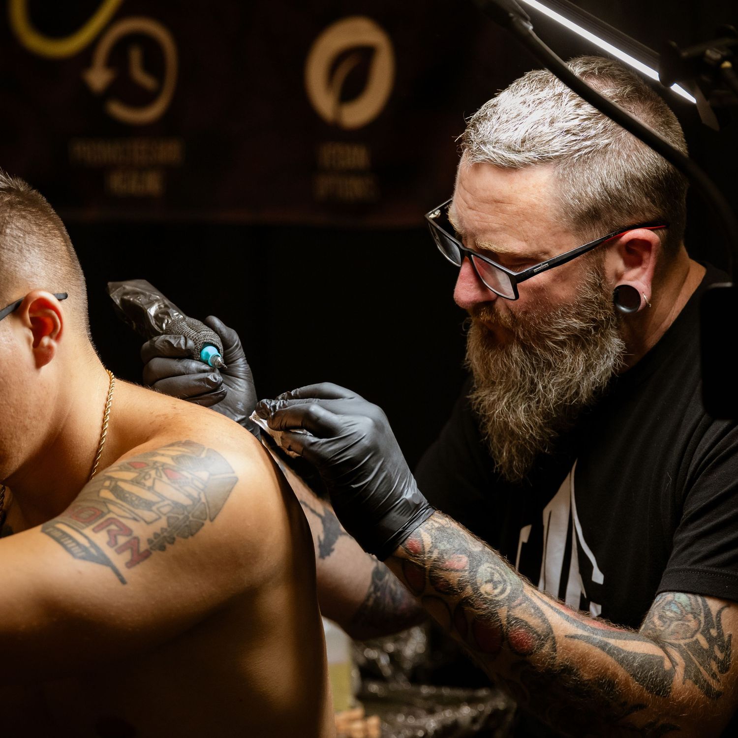 Professional tattoo artist in black clothing and gloves working on a client's shoulder tattoo.