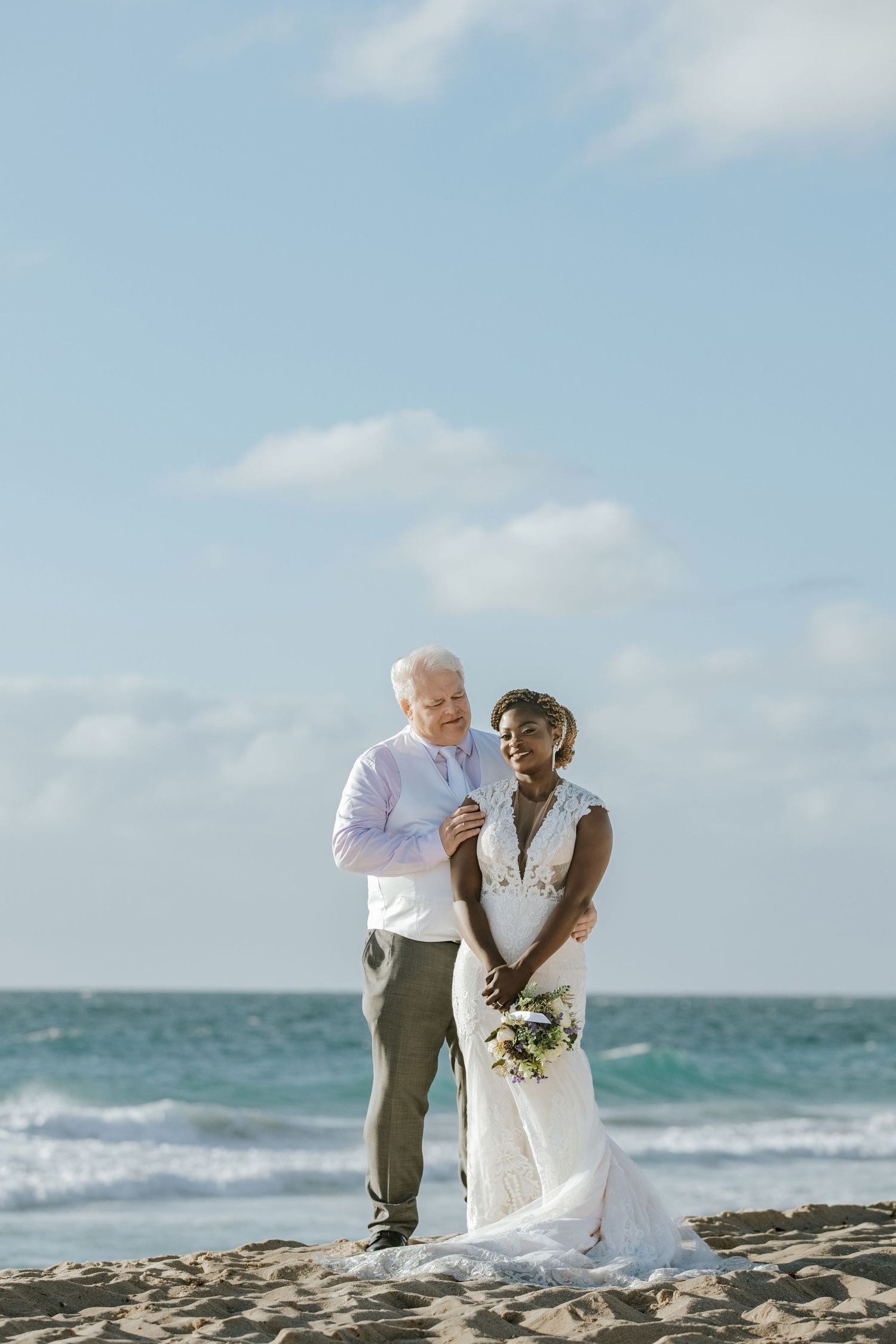 A wedding couple shares a tender moment on a sunny beach with ocean waves in the background.