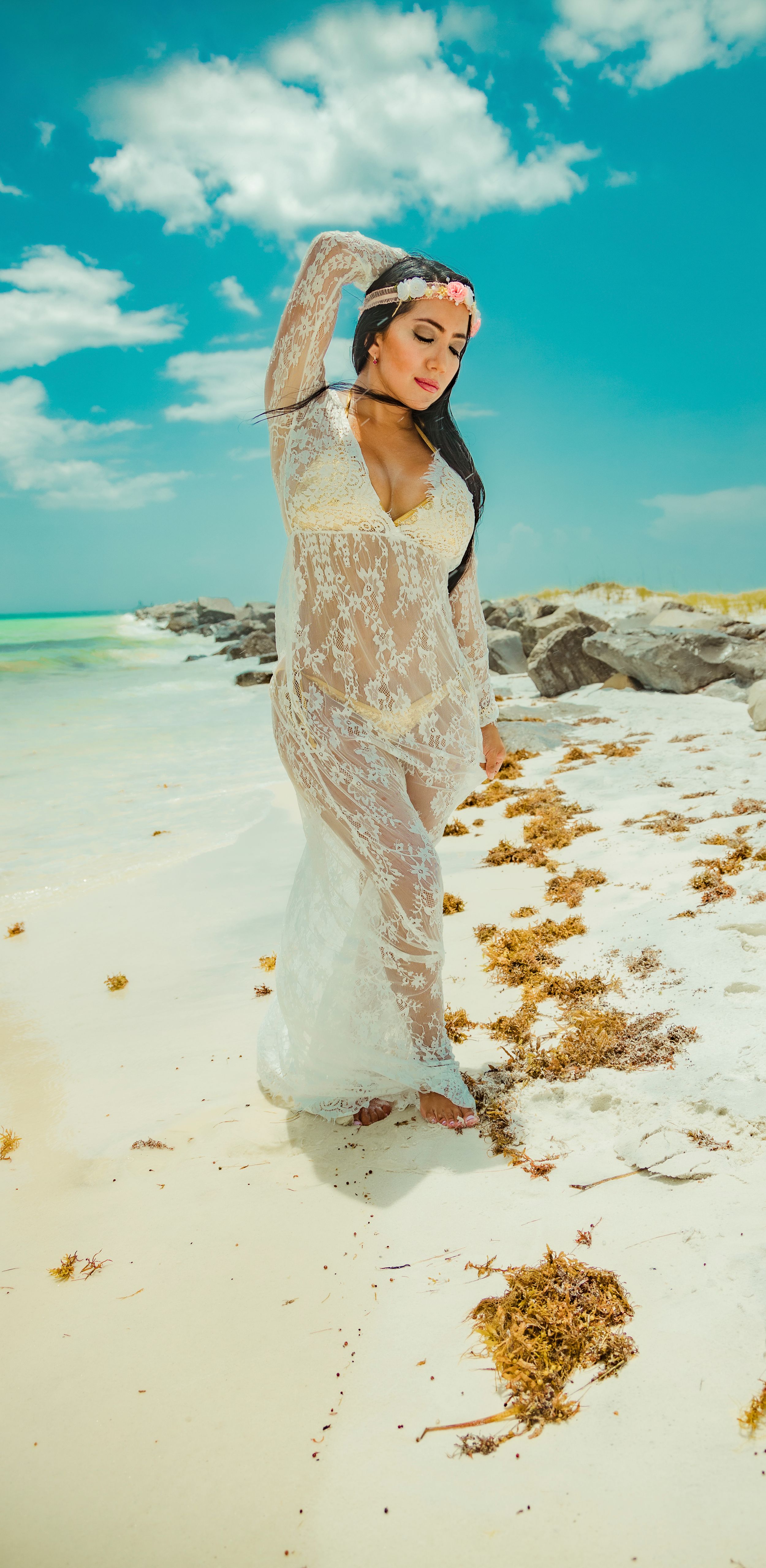 Model in white lace dress poses on sandy beach under vibrant turquoise sky.