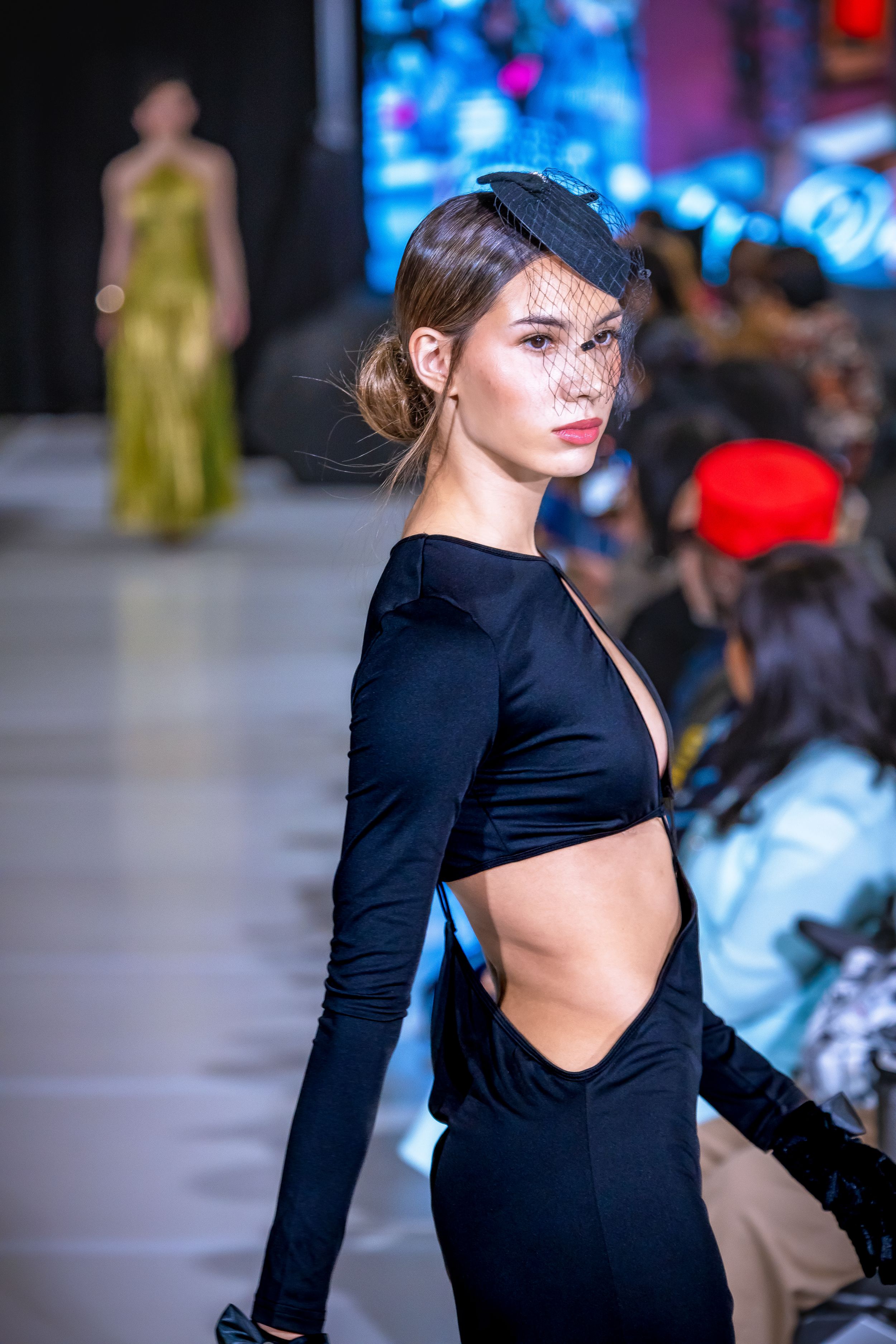 Model walks runway in black cutout dress and veil fascinator at fashion show.