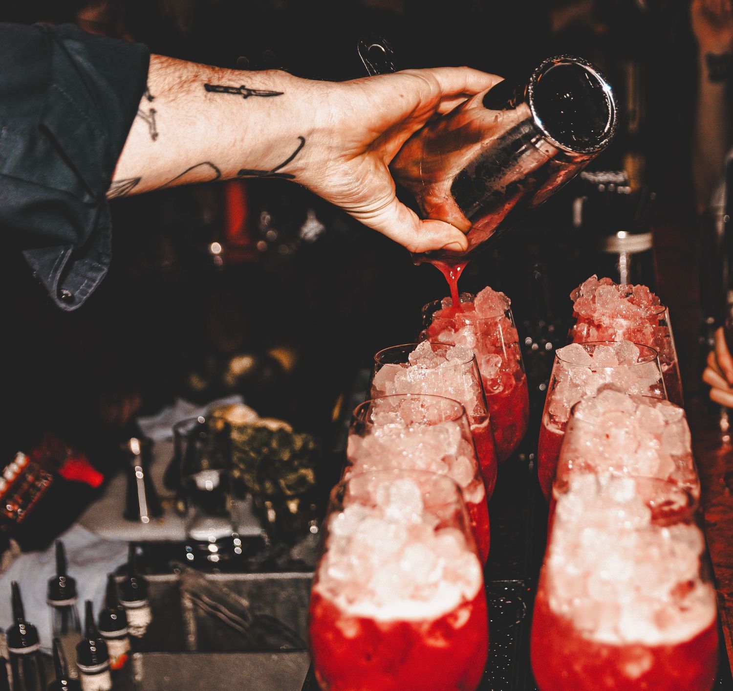 Tattooed hand pouring drinks over crushed ice into cocktail glasses.