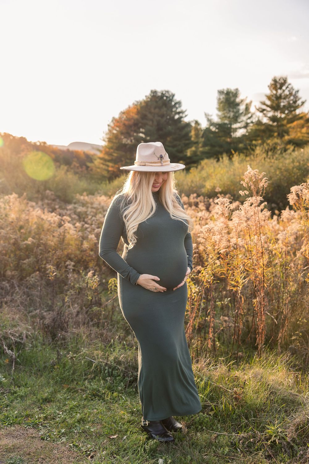 A pregnant woman in a green dress and beige hat stands in a golden autumn field at sunset with tall grass and trees.