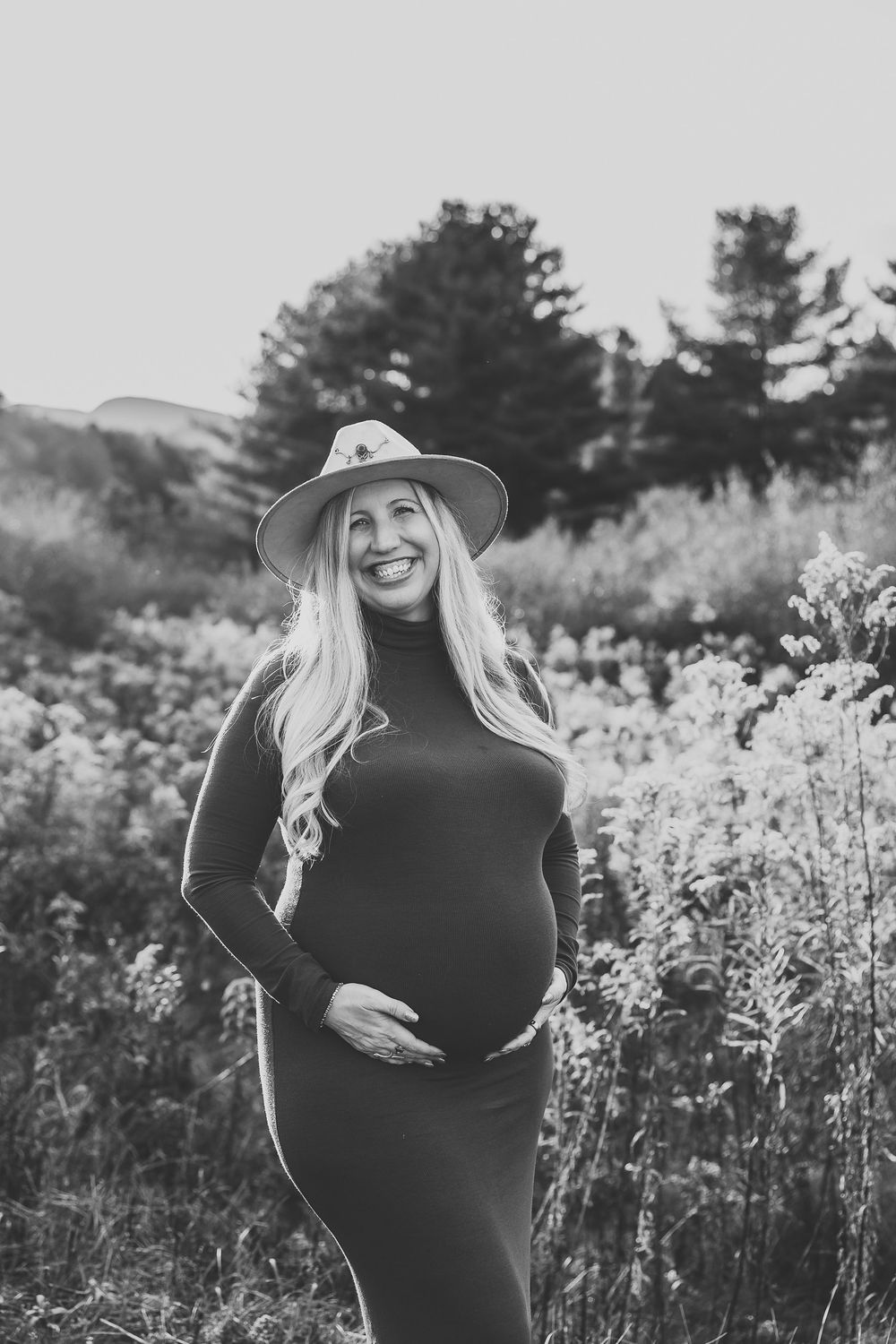 Black and white maternity photo showing a pregnant woman in a dress and hat standing in a wildflower meadow.