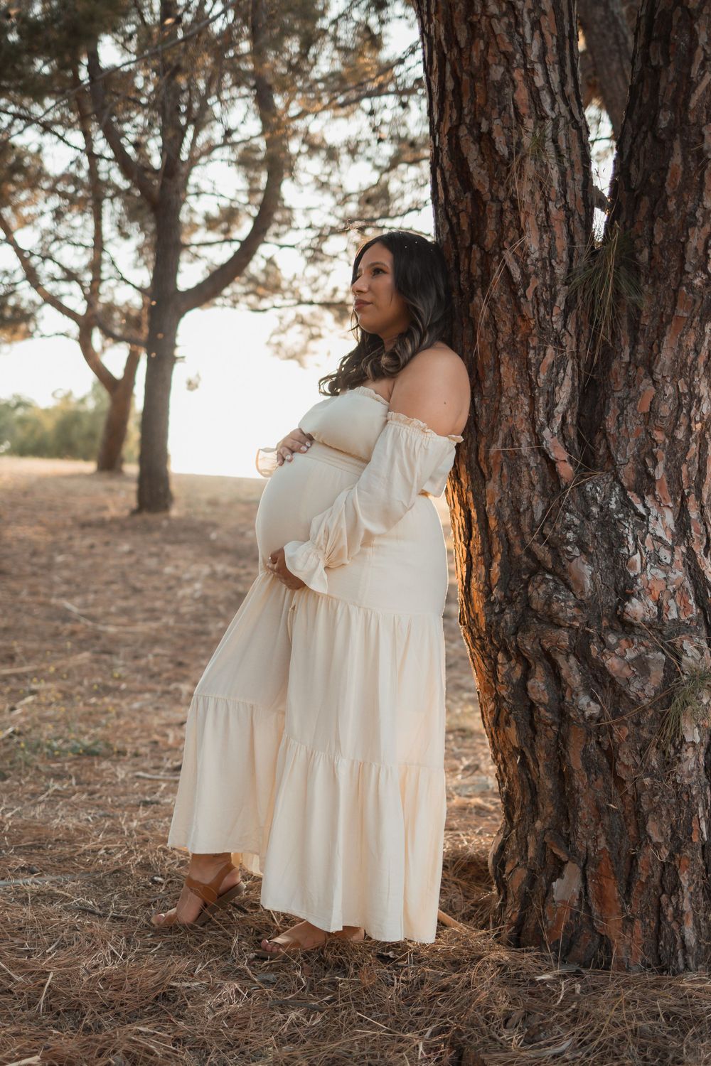 Pregnant woman in flowing white dress poses elegantly against a pine tree.