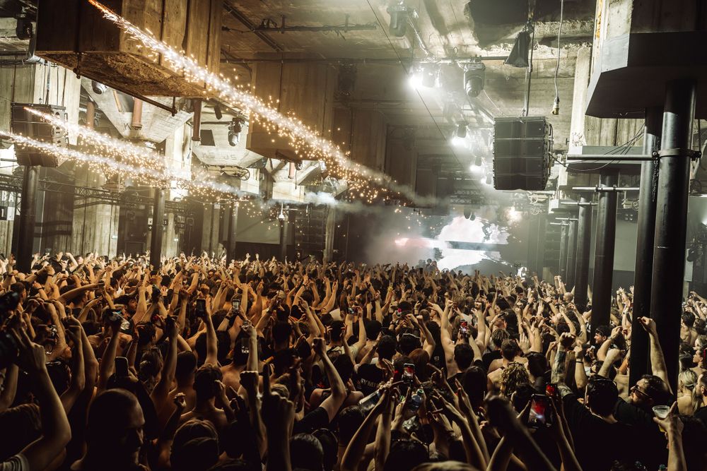 Massive crowd raises hands in unison at an indoor concert venue with bright overhead lighting.