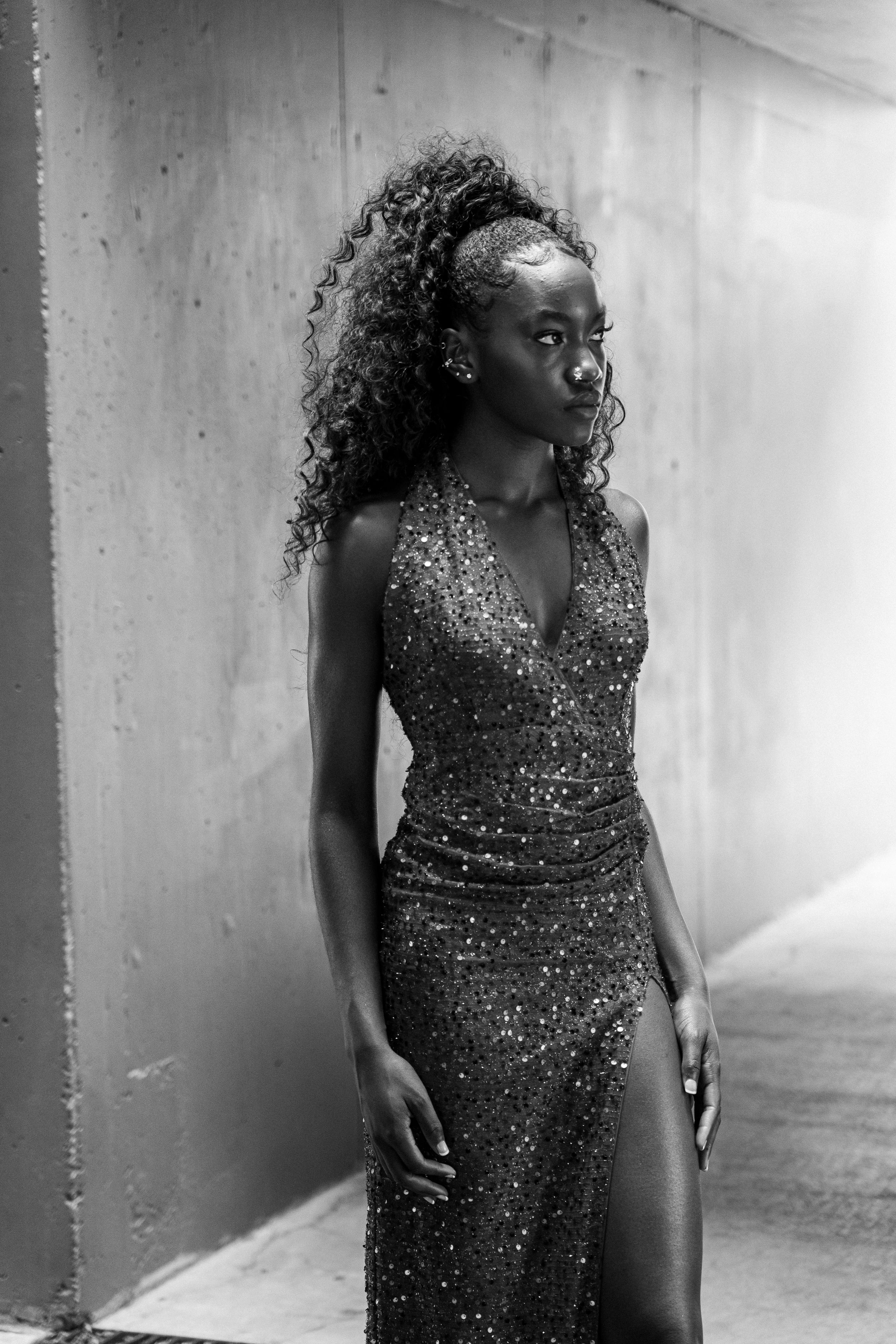 A woman in a sparkly evening gown poses against a concrete wall in black and white photography.