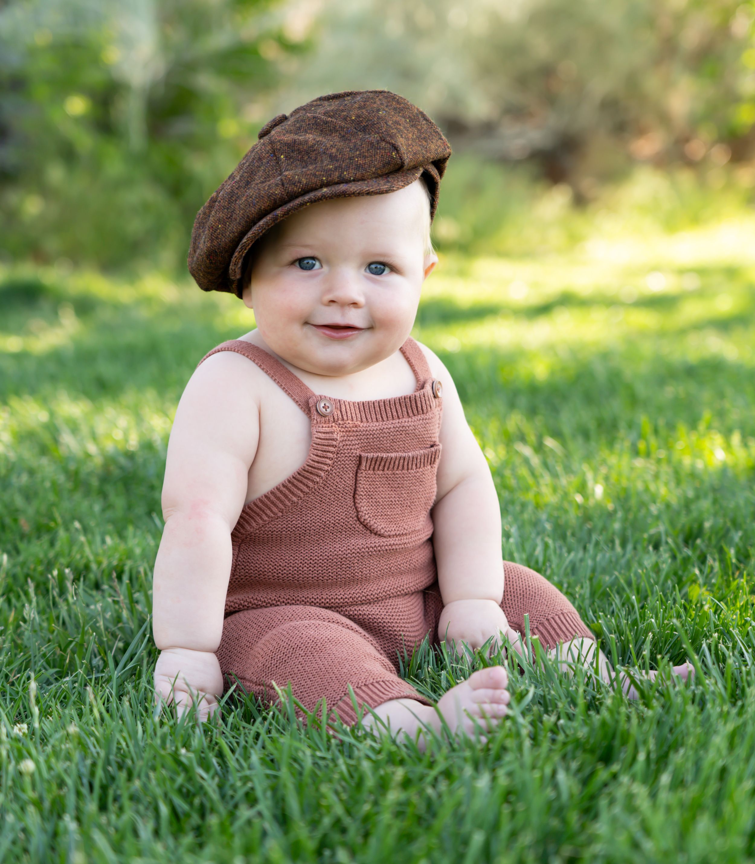 Smiling baby in brown overalls and flat cap sitting on grass in sunlit garden.