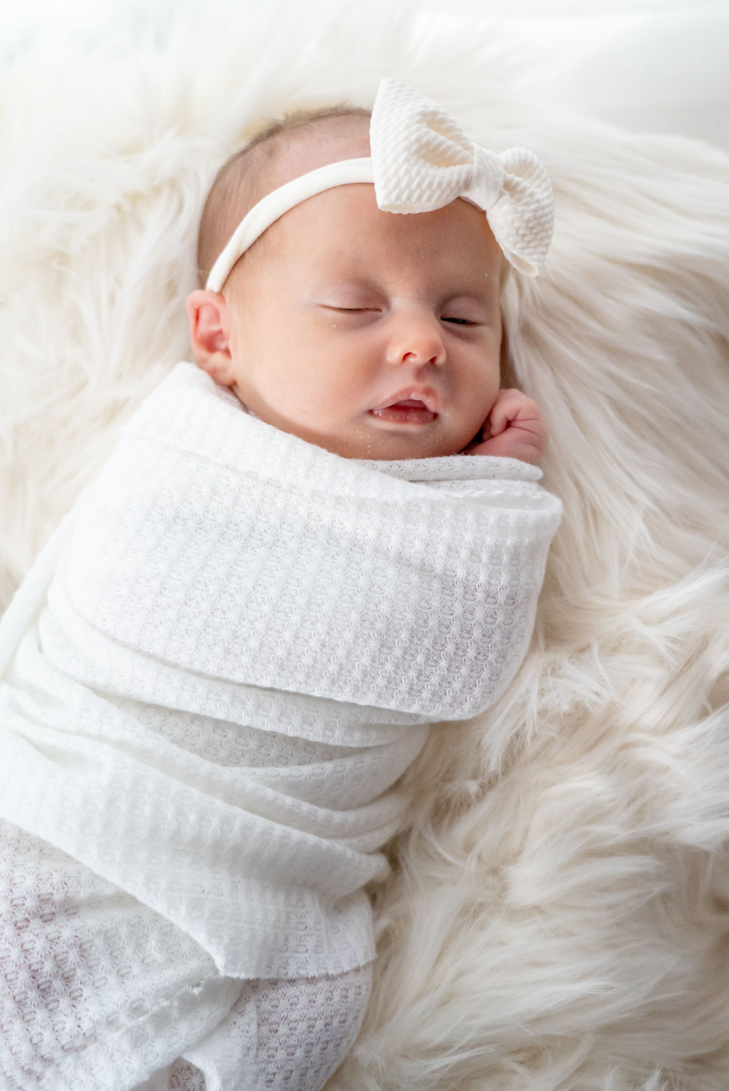 Sleeping newborn wrapped in white swaddle with bow headband on furry blanket.