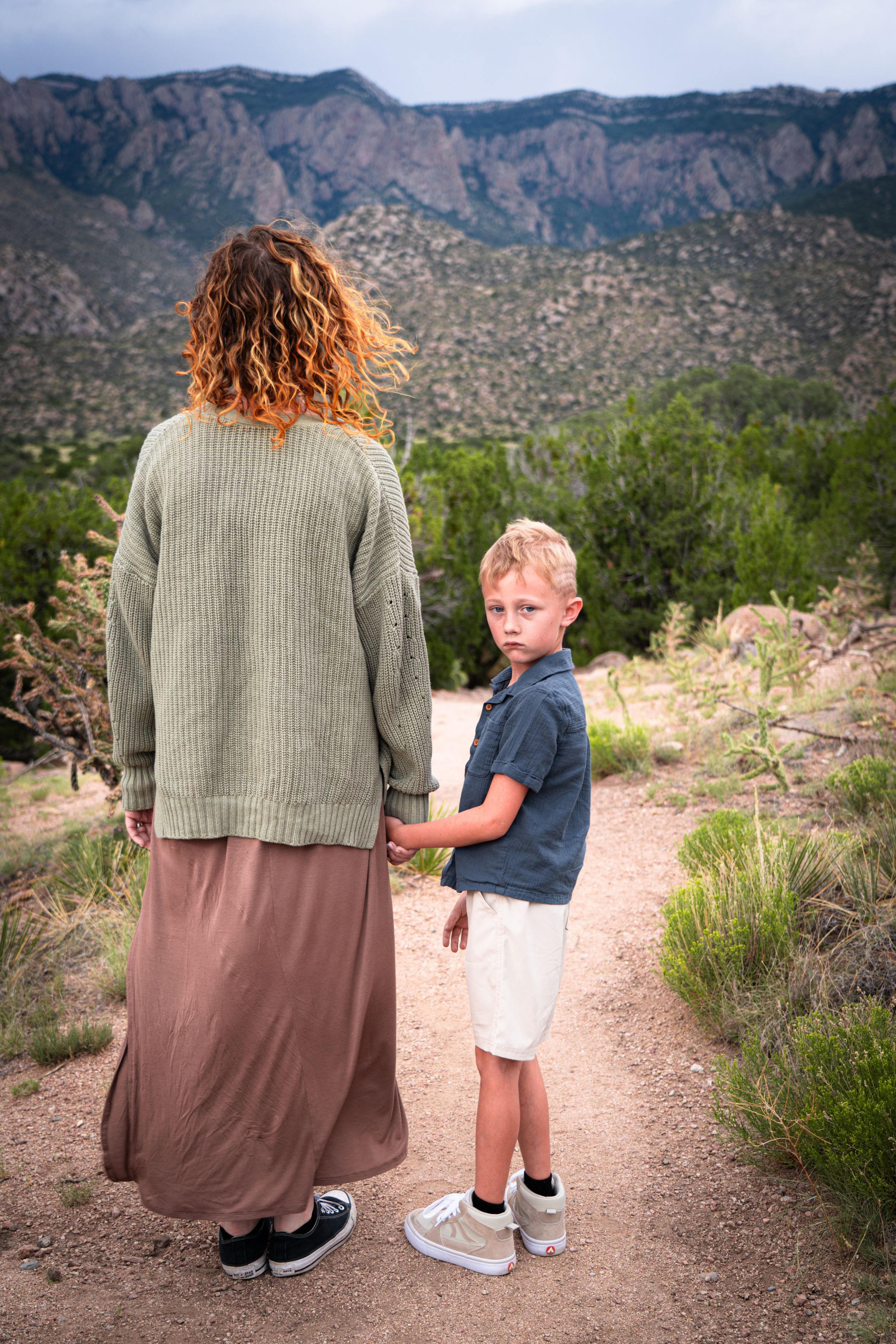 A person and child holding hands while hiking on a desert trail with mountains in the background.