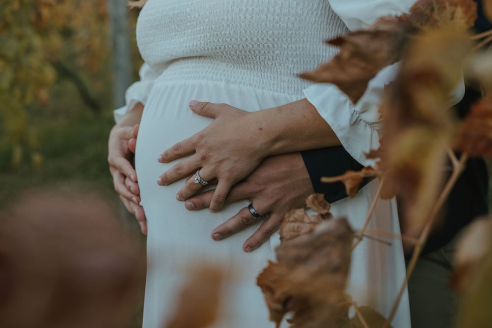 Close up shot of hands cradling a pregnant belly in a white dress against an autumn background.