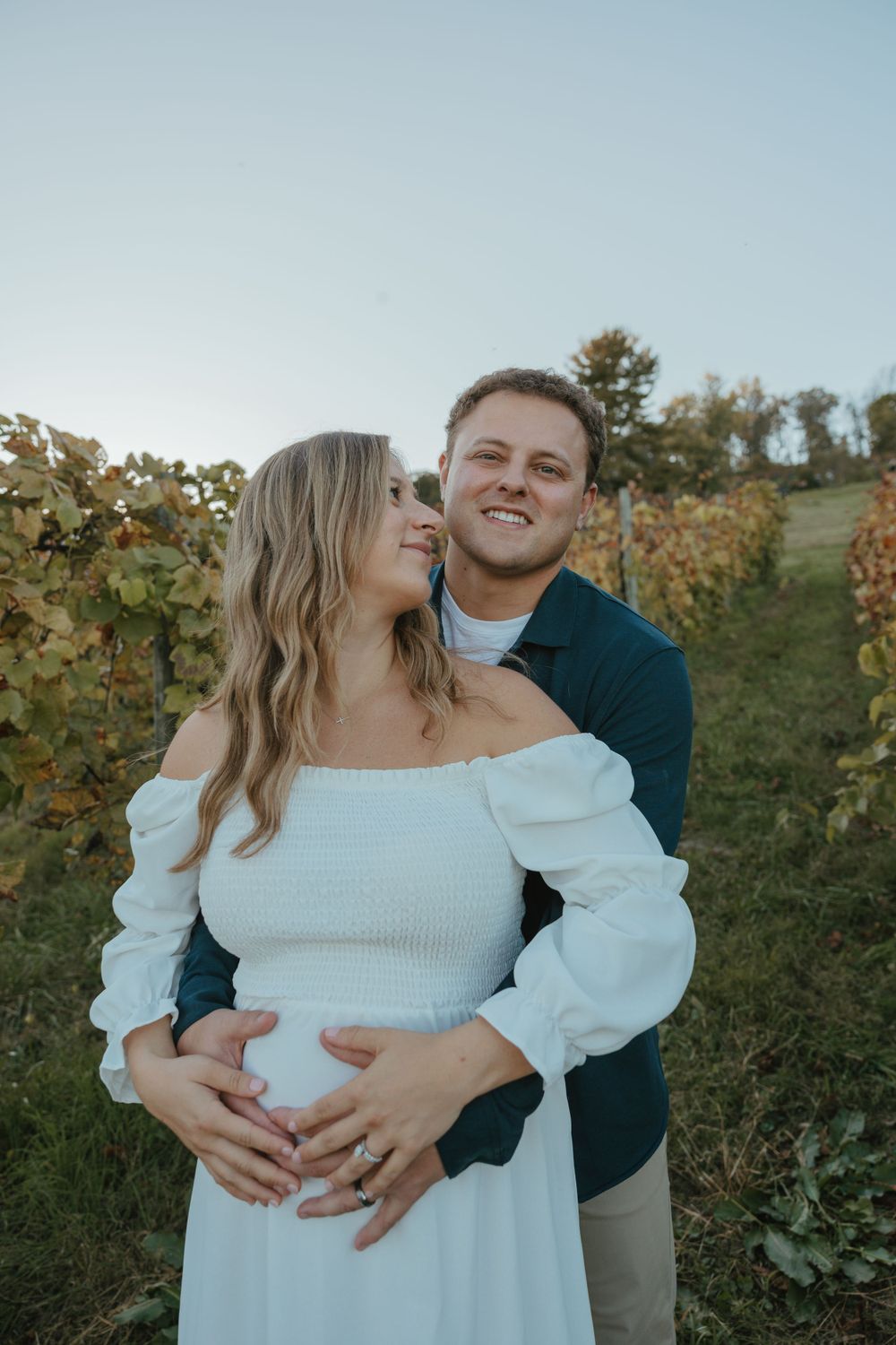 Romantic maternity photo in vineyard with couple in white dress and navy sweater.