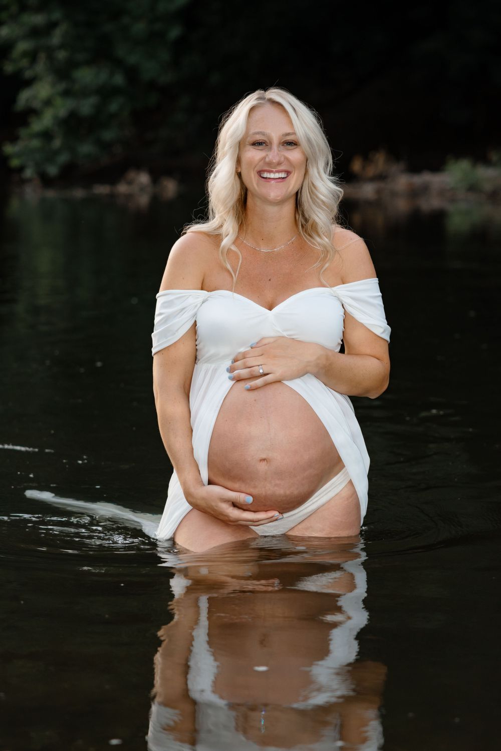Maternity water portrait shows woman in white dress standing in reflective water.