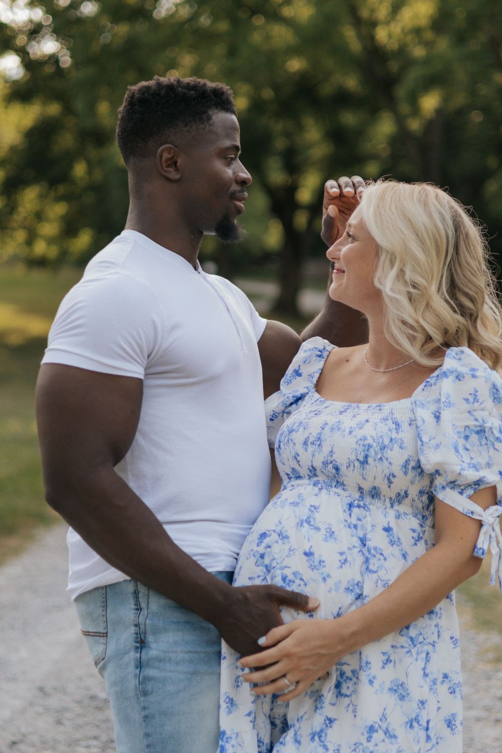 Expecting couple embraces outdoors, woman wearing blue floral dress shows pregnant belly in sunny park setting.