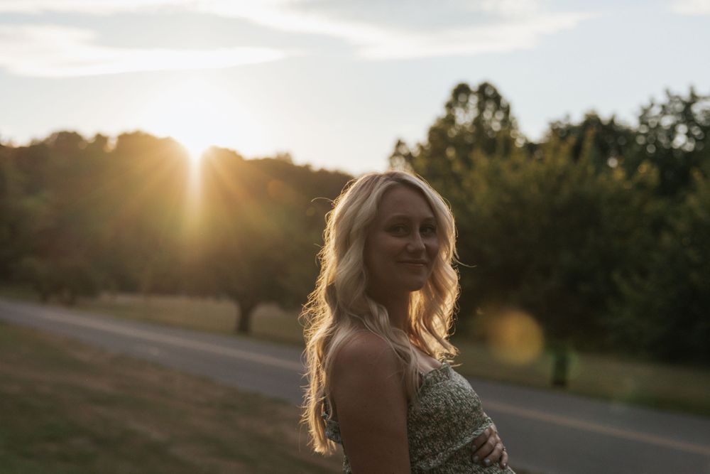 A pregnant woman smiles in a sunlit outdoor setting during golden hour.
