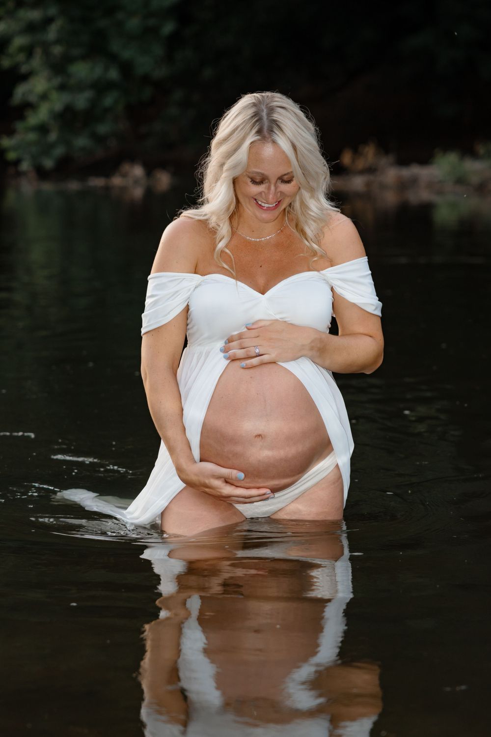Maternity photoshoot in water featuring woman in white off-shoulder dress at sunset.