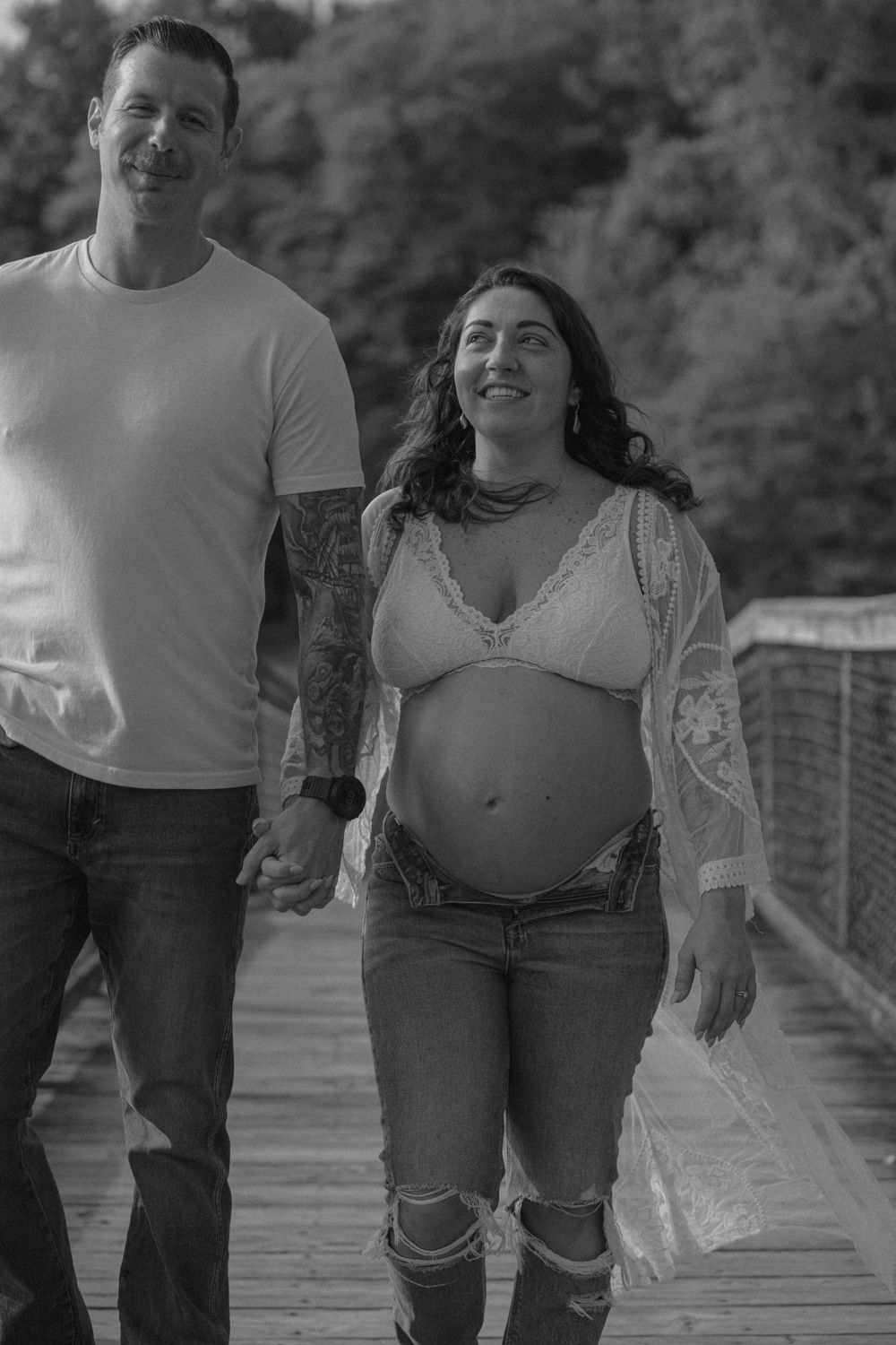 Expectant couple walks together on wooden boardwalk in black and white photo.