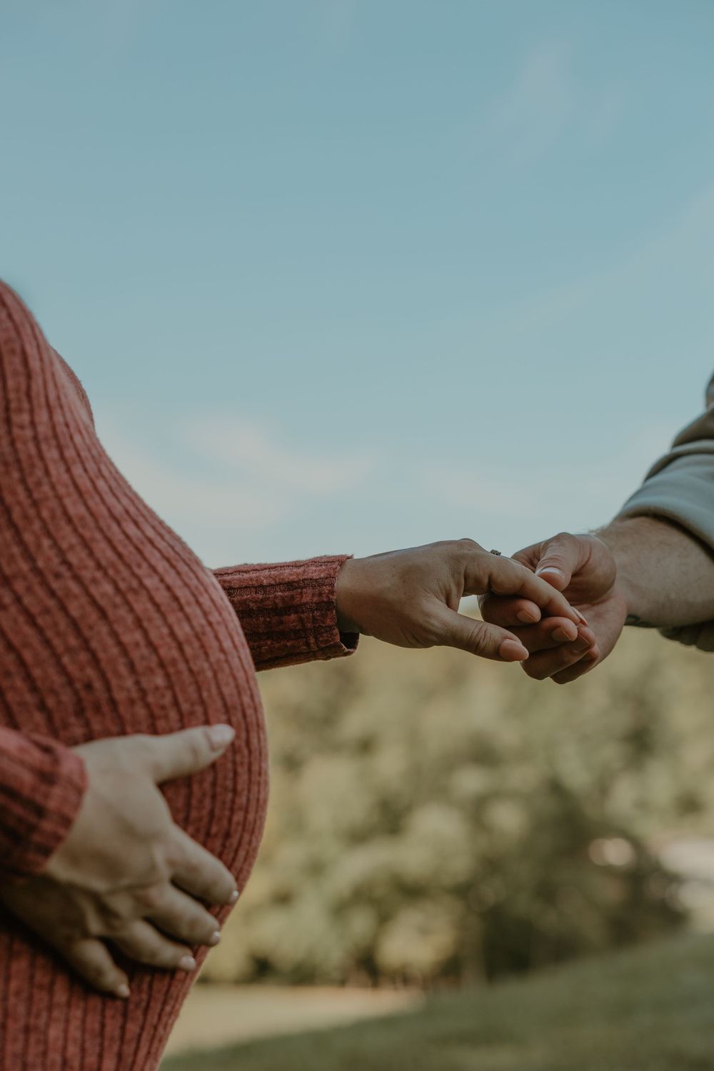 Close-up of hands holding pregnant belly in rustic orange sweater against blue sky.