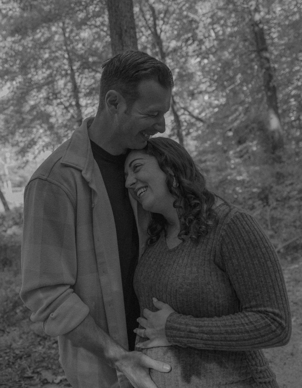 A romantic black and white portrait of a couple embracing in a forest setting.