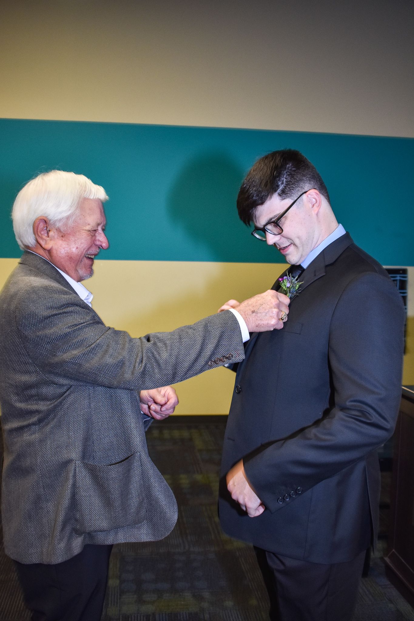 An elderly man helps adjust a boutonniere on a formal black suit.