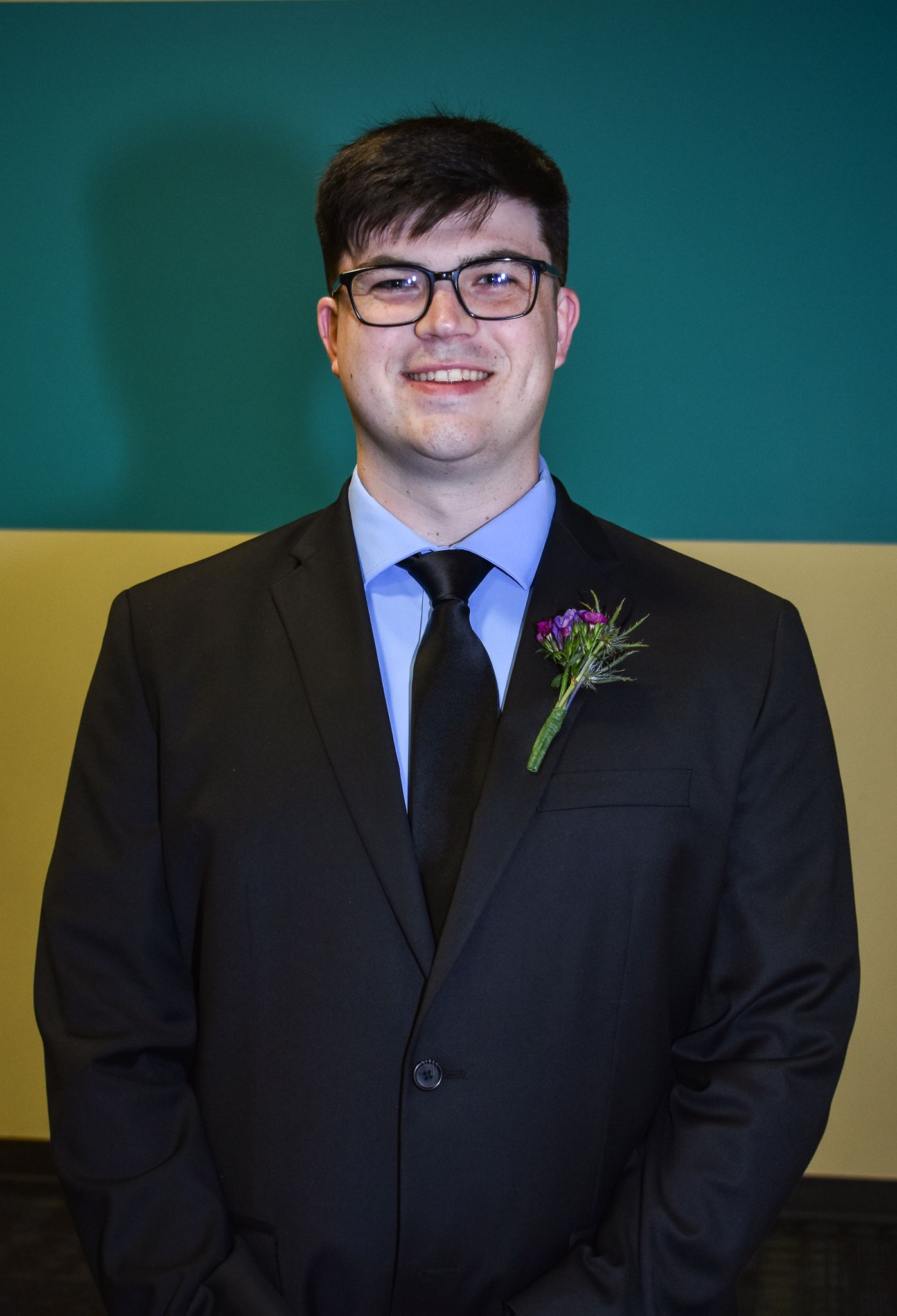 A groom in a dark suit and glasses with a purple floral boutonniere against a teal and yellow wall.