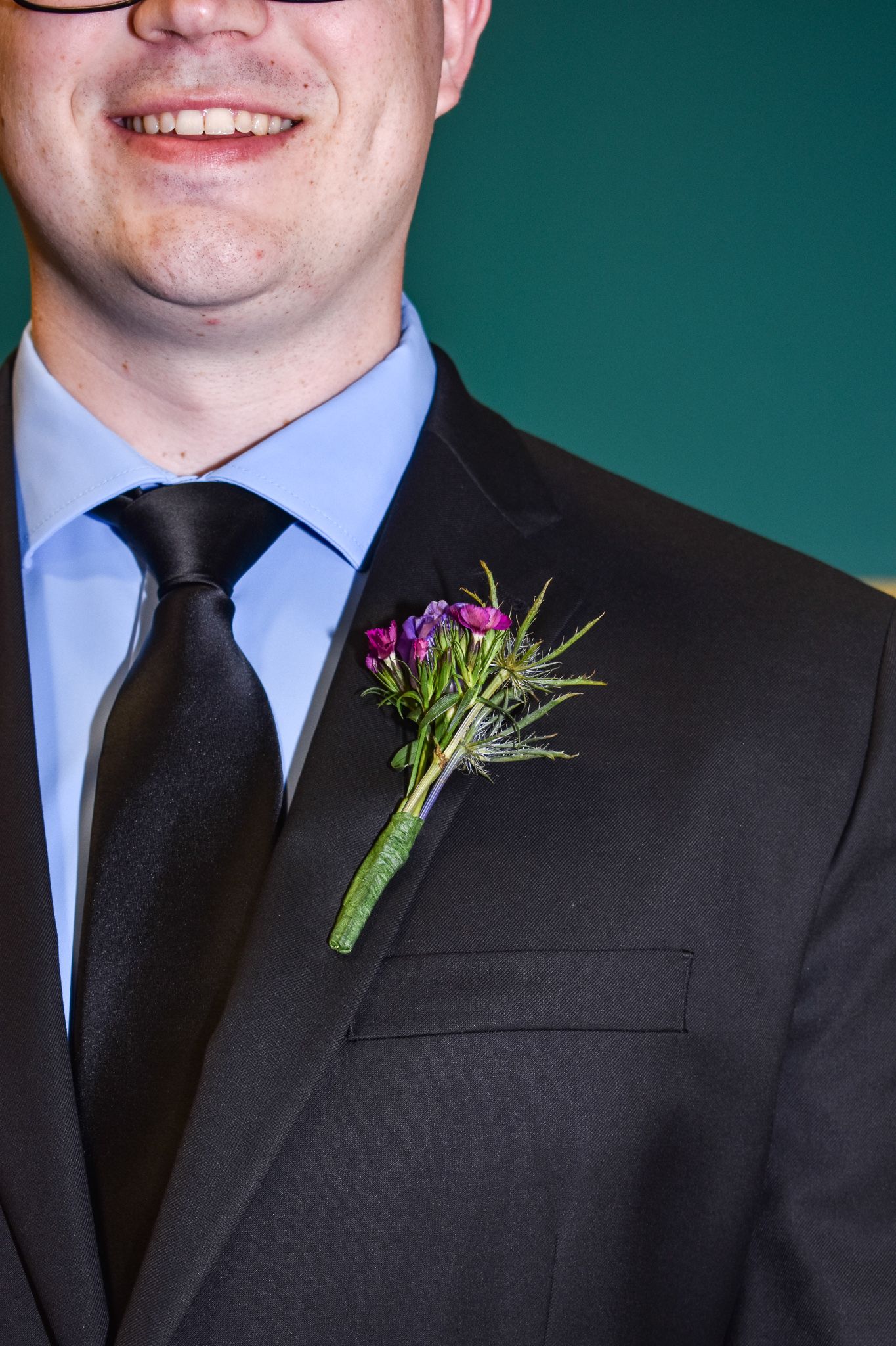 Close-up of a purple floral boutonniere on a black suit jacket with blue shirt and tie.