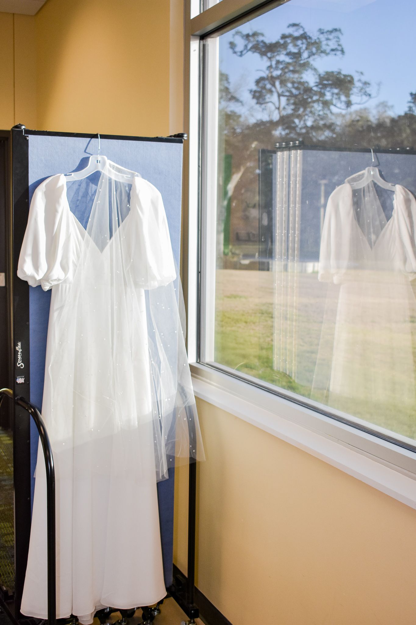 A white wedding dress with short sleeves displayed on a hanger near a sunny window.